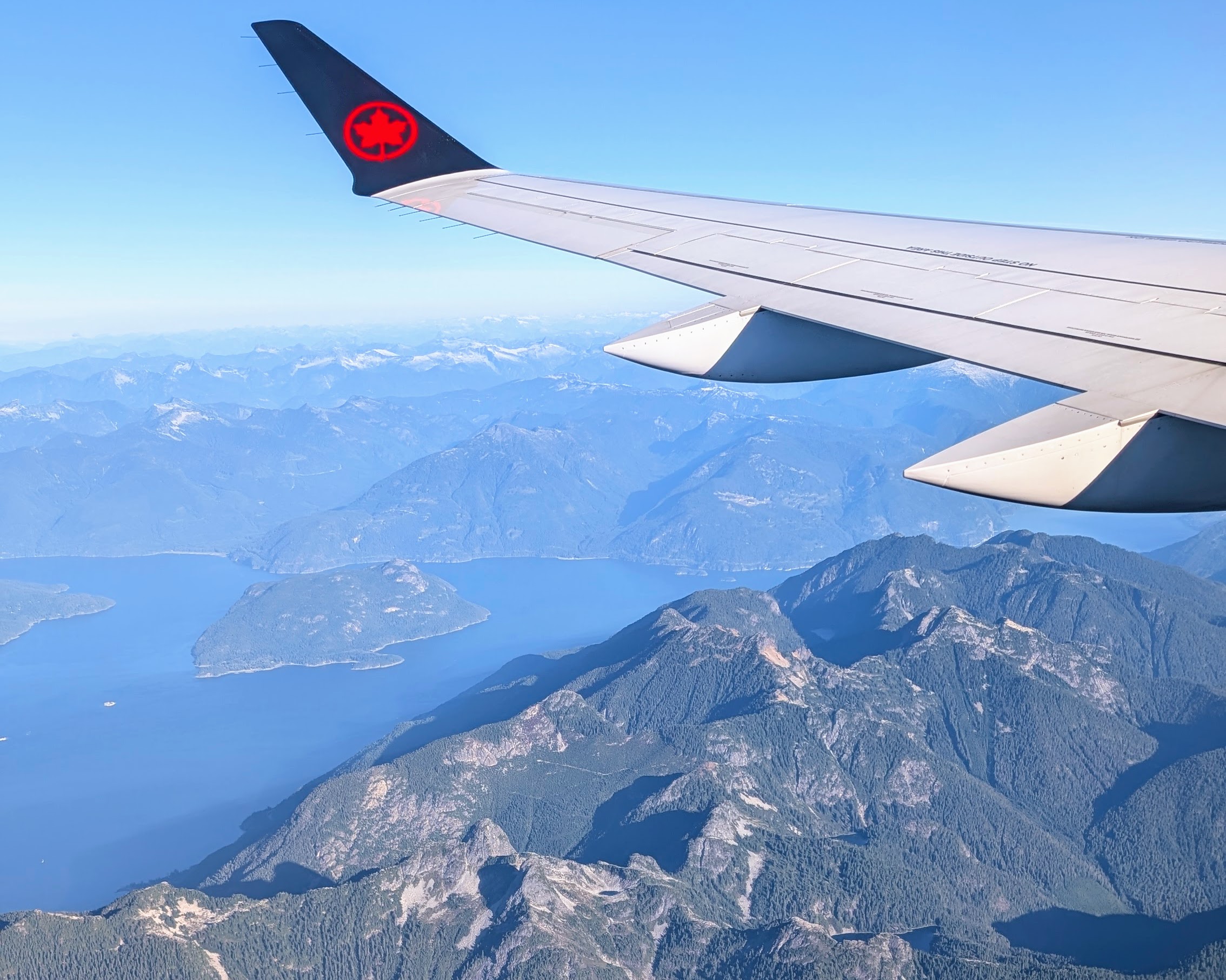 Photo out the window of an airplane over the Pacific ocean and mountains of British Columbia. You can see the wing of the plane and the Air Canada logo at the end of it.