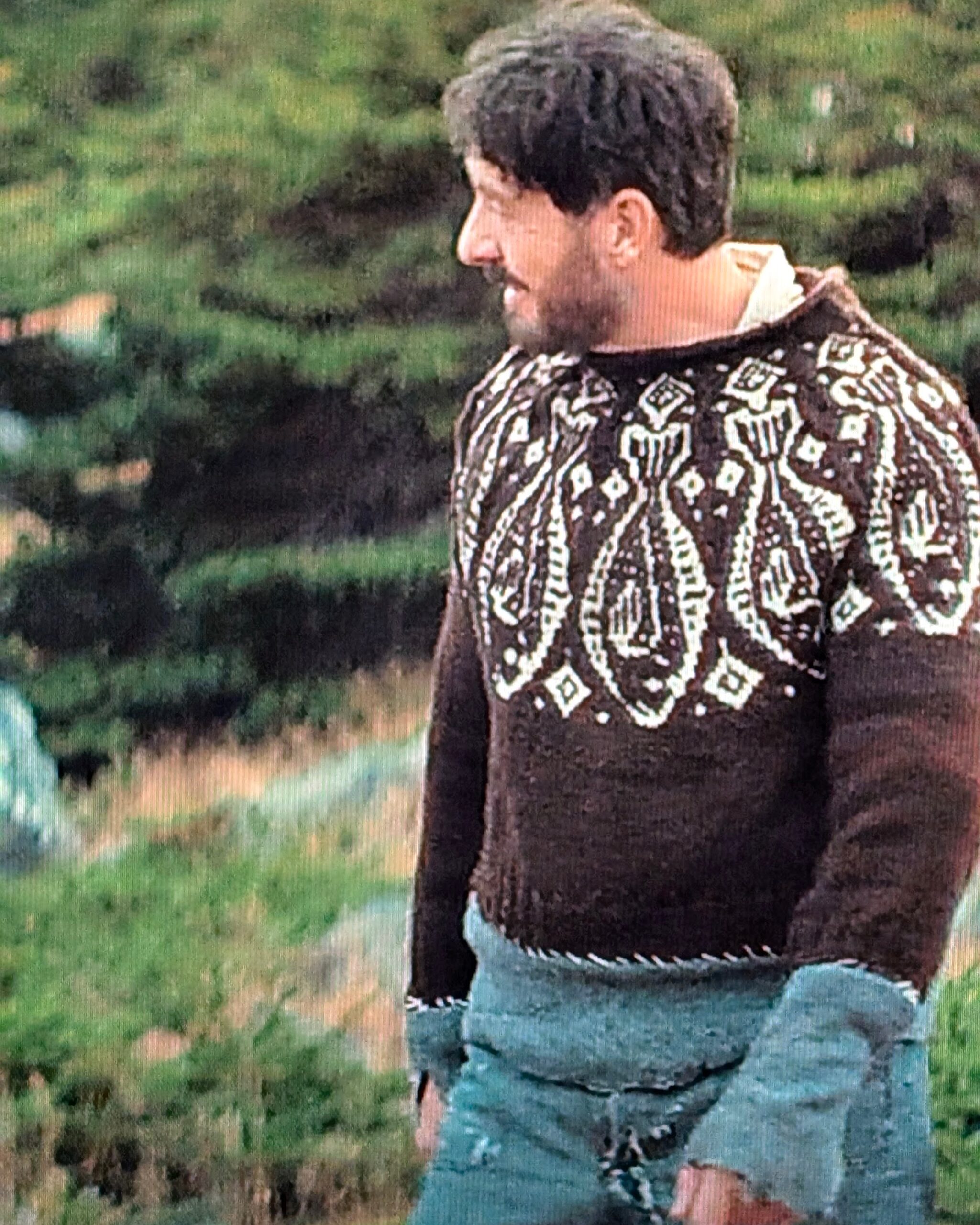 Photo, zoomed in, of my television during an episode of "Below," showing a man in a brown sweater with white fish colourwork around the yoke, with several inches of a different knitted fabric visibly whipstitched to the bottom hem and sleeve cuffs.